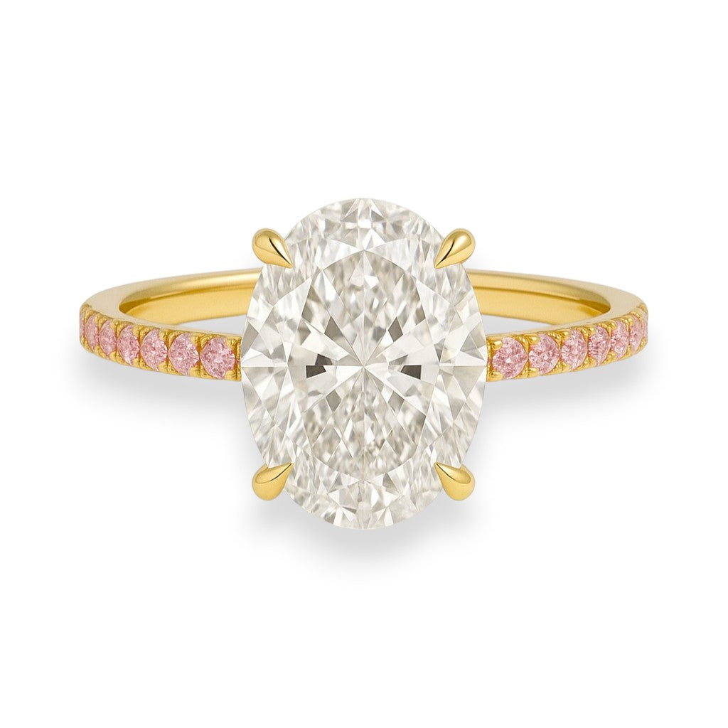 Gold ring with a large diamond and smaller pink diamonds on the band against a white background. his ring is a dream in colour and sparkle. Featuring a 4.7ct oval crushed ice moissanite centre stone and a band lined with delicate pink sapphires, it's a fresh, romantic take on the classic solitaire — made for the bride who loves a hint of softness with her brilliance.