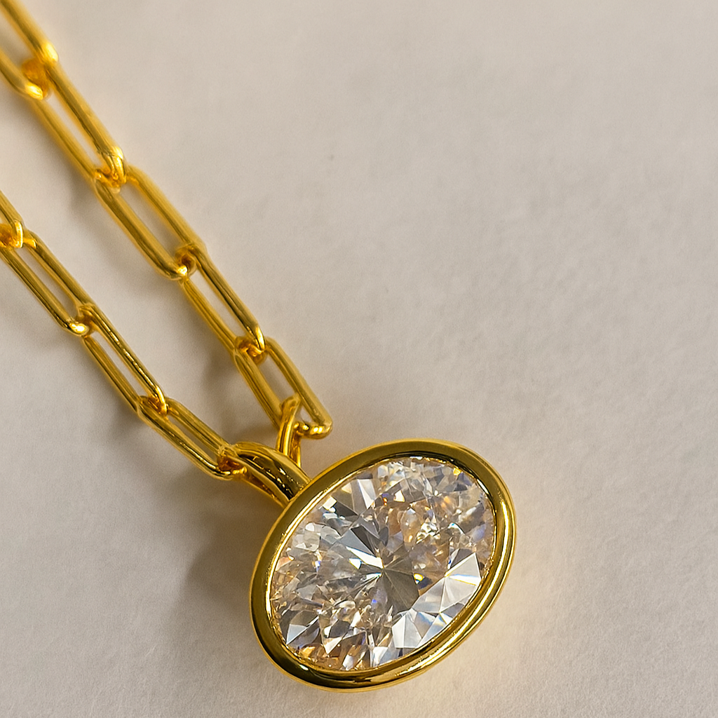 Gold necklace with a large diamond pendant on a light background. 6ct oval cut moissanite bezel pendant with yellow gold attachable bail on a paper clip chain
