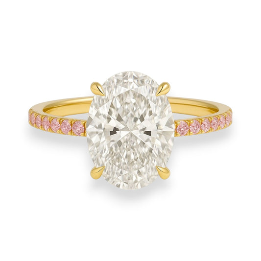 Gold ring with a large diamond and smaller pink diamonds on the band against a white background. his ring is a dream in colour and sparkle. Featuring a 4.7ct oval crushed ice moissanite centre stone and a band lined with delicate pink sapphires, it's a fresh, romantic take on the classic solitaire — made for the bride who loves a hint of softness with her brilliance.