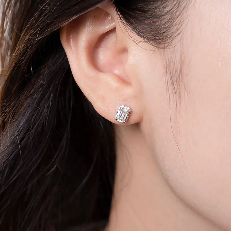 Close up of brunettes ears wearing a 1 carat emerald cut moissanite stud with halo in14K gold